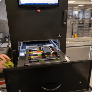 Un iPhone usagé est coincé sur des carreaux colorés dans un dispositif carré. À côté, on peut voir deux mains tenant un chiffon de nettoyage.