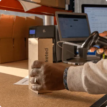 Une main gauche pose une boîte en carton contenant le smartphone entièrement rénové sur une table